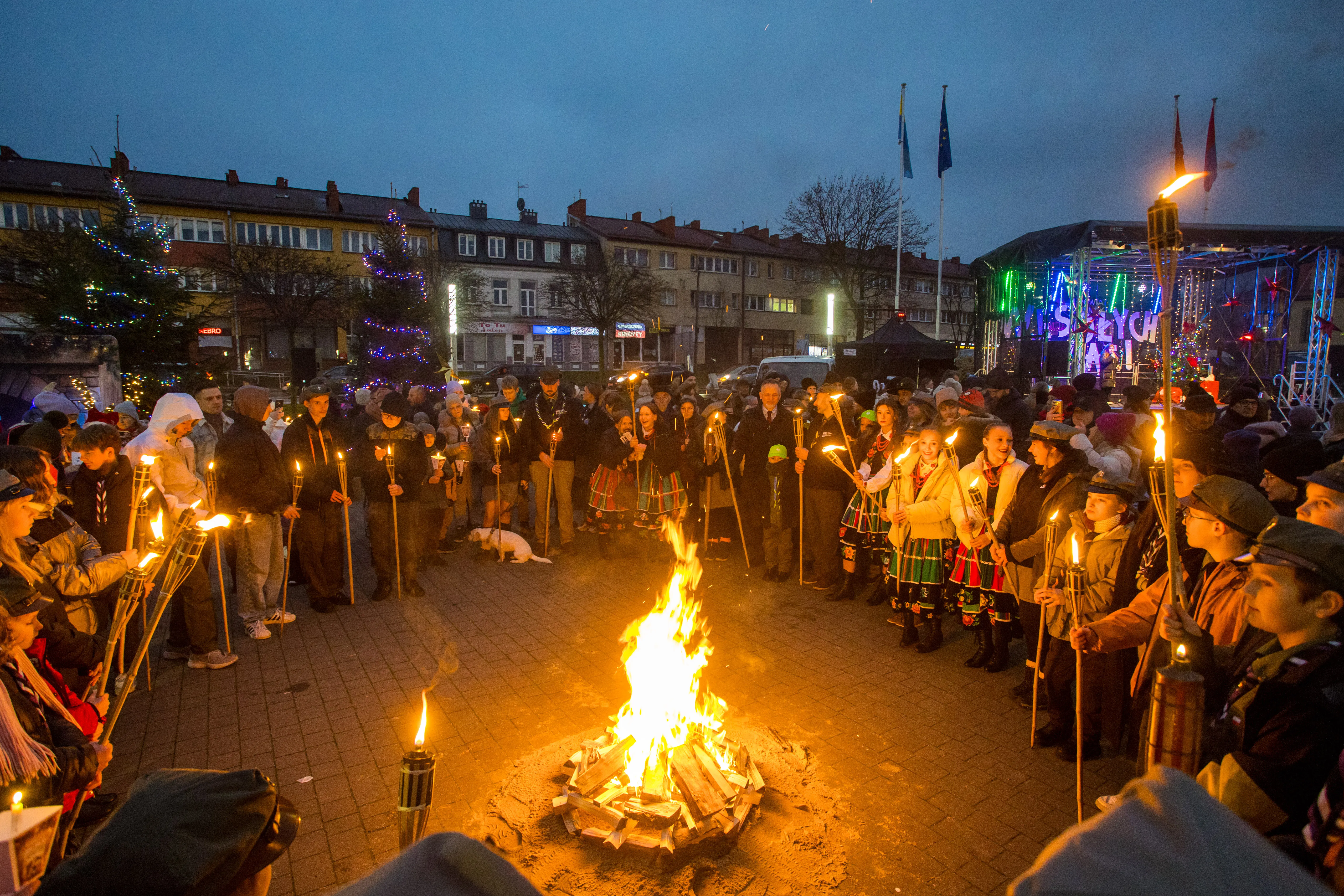 Ludzie stojący w kręgu na rynku wieczorem, trzymający pochodnie, otaczają palące się ognisko, w tle oświetlone budynki i scena z dekoracjami świątecznymi.